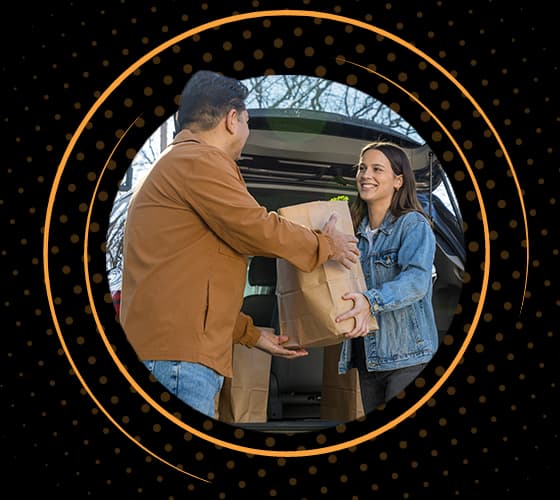 A smiling woman handing a bag of groceries to a man in a brown jacket from the back of an SUV.